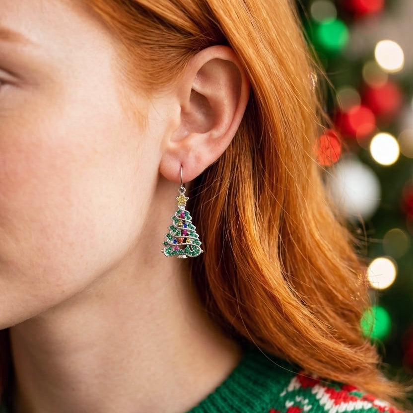 A close-up of a woman with red hair wearing a dangling Christmas tree earring. The earring features a silver-tone hook and frame set with green, red, and blue cubic zirconia crystals, topped with a small gold star.