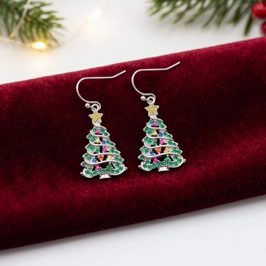 A pair of silver-toned dangle earrings shaped like Christmas trees, decorated with green, pink, purple, and gold cubic zirconia crystals, resting on a deep red velvet surface with festive pine branches in the background.