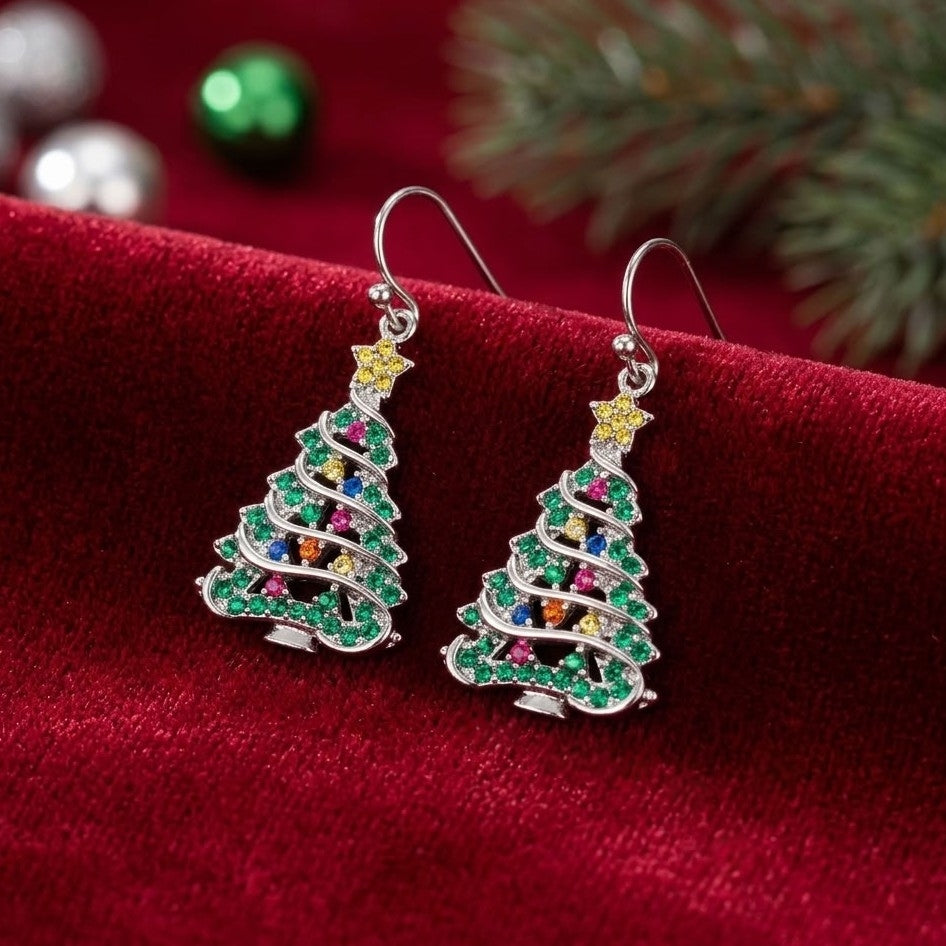 A pair of silver tree-shaped drop earrings embellished with green, pink, blue, orange, and yellow cubic zirconia crystals, resting on a red velvet surface with festive holiday decorations in the background.