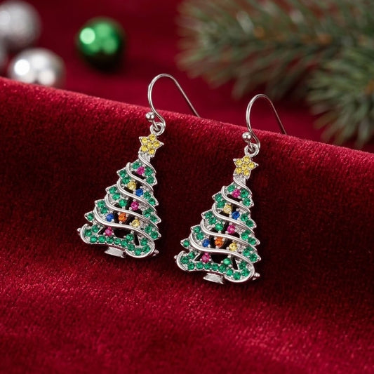 A pair of silver tree-shaped drop earrings embellished with green, pink, blue, orange, and yellow cubic zirconia crystals, resting on a red velvet surface with festive holiday decorations in the background.