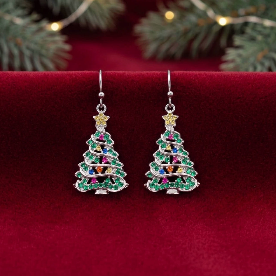 A pair of silver-toned dangle earrings shaped like Christmas trees, decorated with green, red, blue, and purple cubic zirconia crystals and topped with gold star-shaped accents, displayed on a red velvet surface with festive greenery and warm lights blurred in the background.