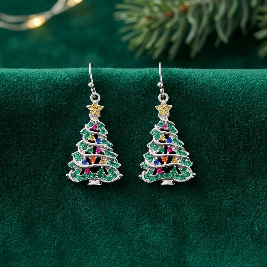 A pair of silver Christmas tree drop earrings embellished with green cubic zirconia crystals and small multicolor gem accents, displayed on a dark green velvet surface with blurred pine branches and warm string lights in the background.