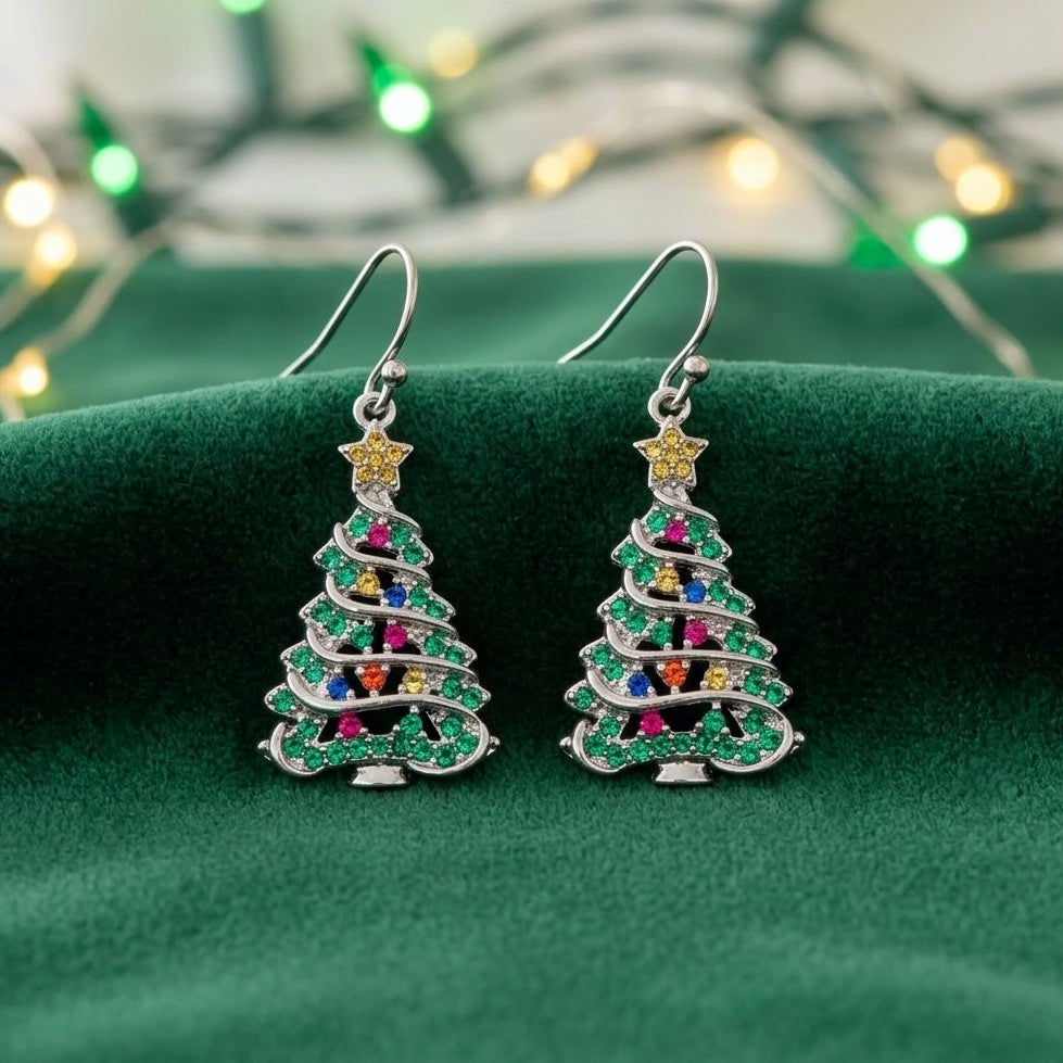 A pair of silver-toned dangle earrings shaped like Christmas trees, decorated with green cubic zirconia crystals and small multicolored gem accents, displayed on a dark green velvet surface with blurred holiday lights in the background.