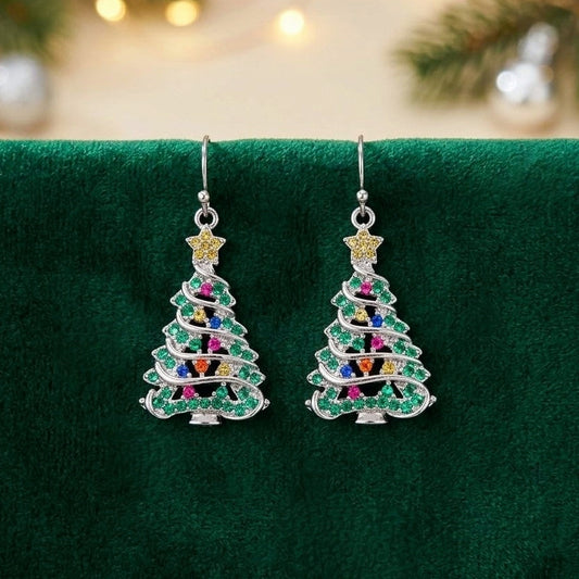 A pair of silver-tone dangle earrings shaped like Christmas trees, decorated with green cubic zirconia crystals and small multicolor gem accents, displayed on a dark green velvet surface with blurred holiday lights in the background.
