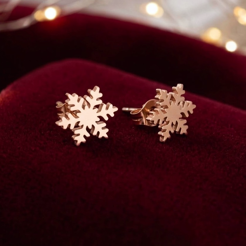 A pair of rose gold snowflake stud earrings displayed on a dark red velvet jewelry box with warm bokeh lights in the background.