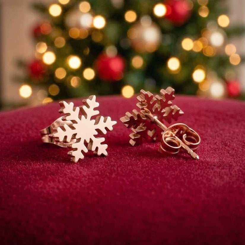 A pair of rose gold snowflake-shaped earrings displayed on a deep red velvet surface, with a blurred Christmas tree in the background.