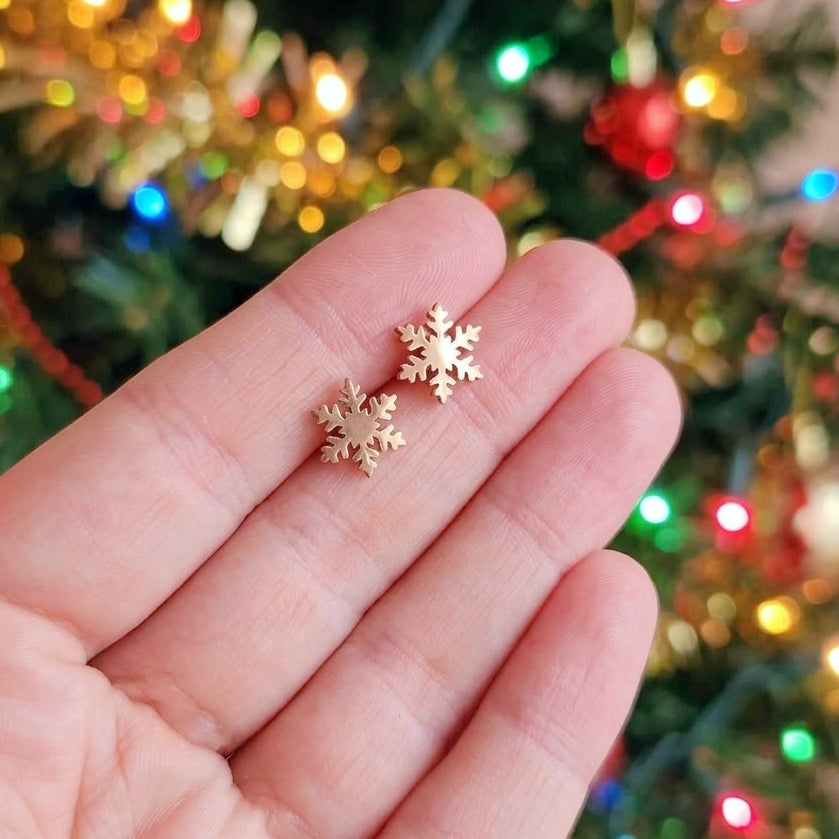 Two small rose gold snowflake stud earrings held on a person's fingers, with a blurred Christmas tree and colorful lights in the background.