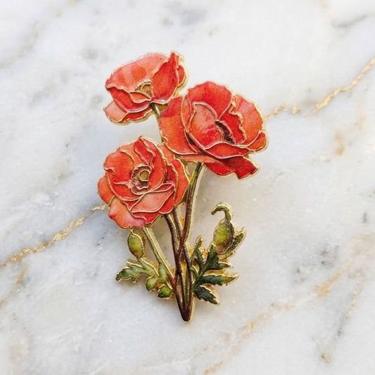 A small floral enamel pin featuring three coral-red poppy-like flowers with green leaves and stems, placed on a white marble surface with faint gold veining.