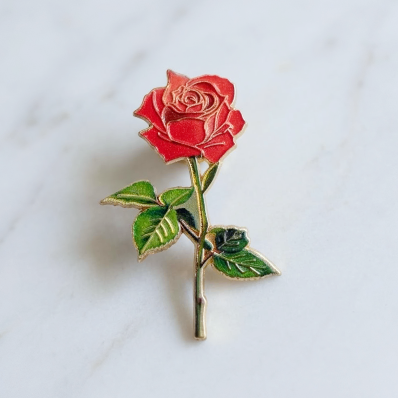 A single rose-shaped enamel pin with a red bloom, green leaves, and a gold-toned stem, placed on a light marble surface.