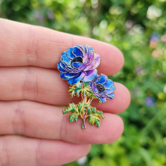 A close-up of a hand holding a small enamel flower pin with two blue and purple poppy or anemone-like blooms and green leaves, against a blurred green outdoor background.