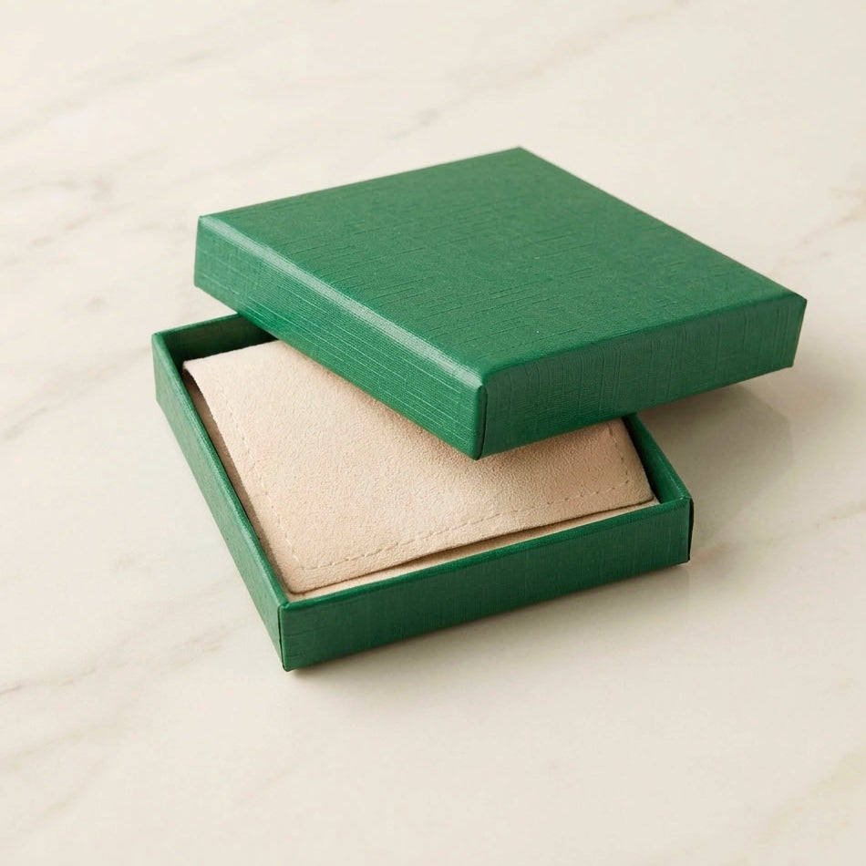 A square green presentation box with its lid propped open, revealing a beige textured pouch inside, set on a light marble-like surface.
