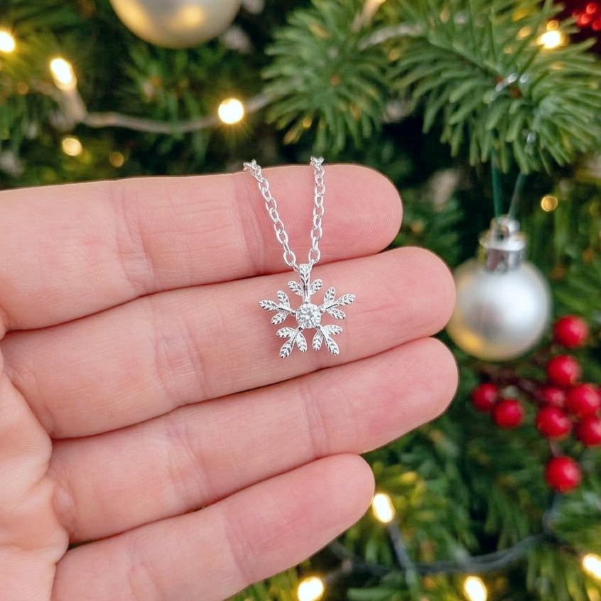 A silver snowflake pendant necklace with a clear central gemstone, held in a person's hand in front of a blurred Christmas tree background with lights and ornaments.