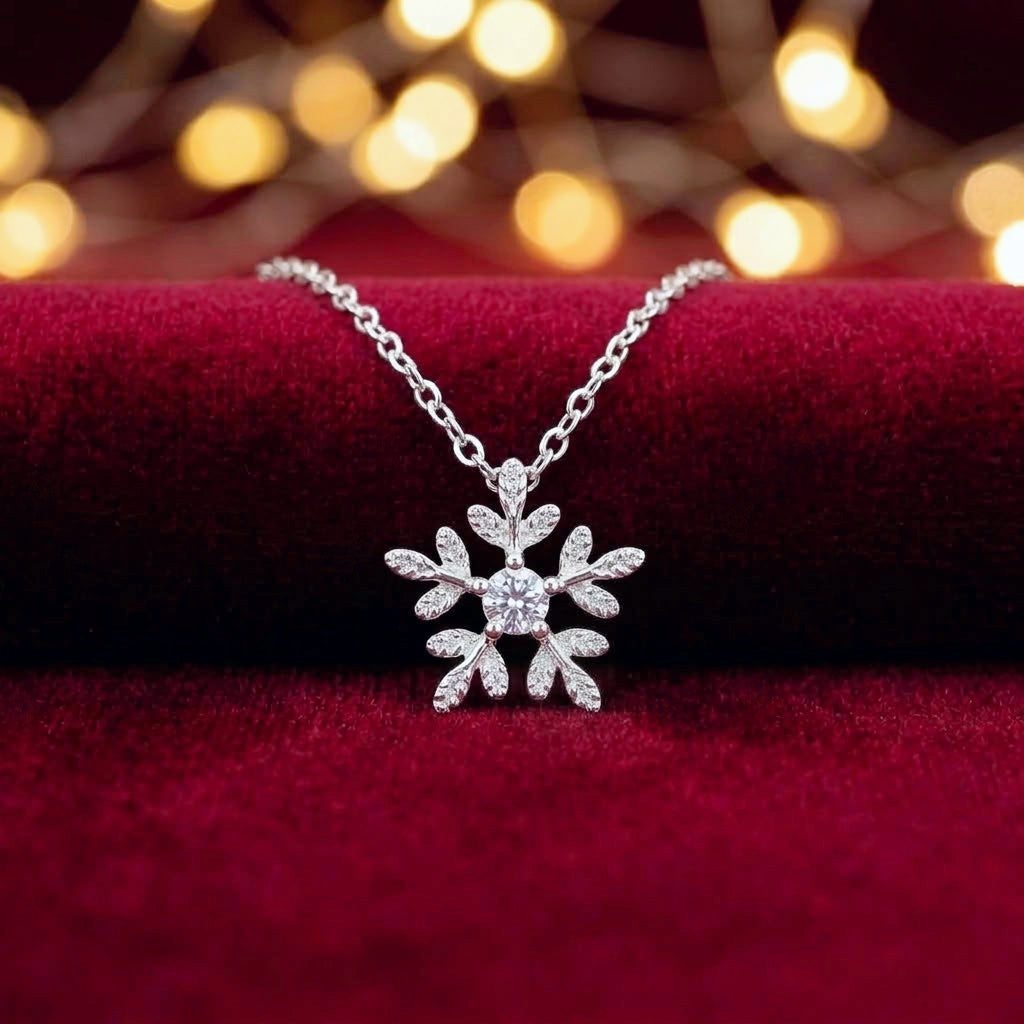 A silver snowflake pendant necklace with a sparkling clear stone, displayed on a deep red velvet surface with warm golden bokeh lights in the background.