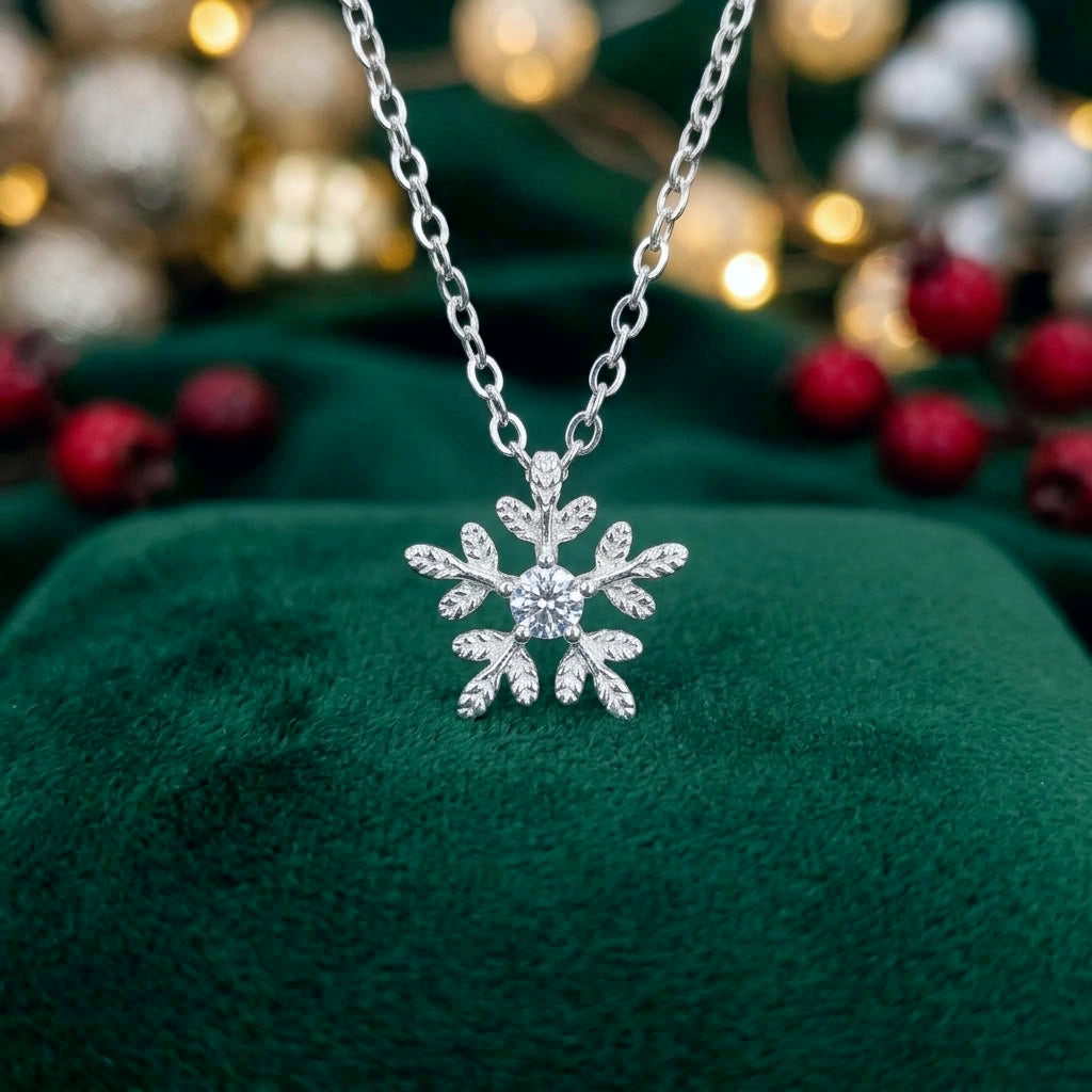 A silver snowflake pendant necklace with a sparkling clear stone, displayed on a deep green velvet surface with warm golden bokeh lights in the background.