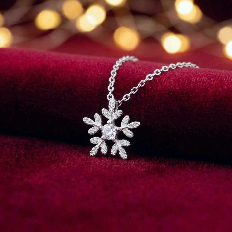 A silver snowflake pendant necklace with a sparkling clear stone, displayed on a deep red velvet surface with warm golden bokeh lights in the background.