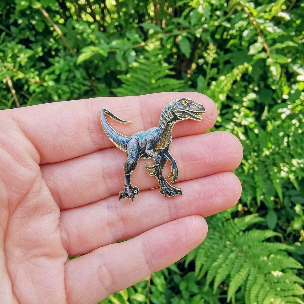 Three small dinosaur pins arranged side by side on a light gray surface: a brown t-rex on the left, a blue raptor dinosaur in the center, and a gold triceratops on the right.