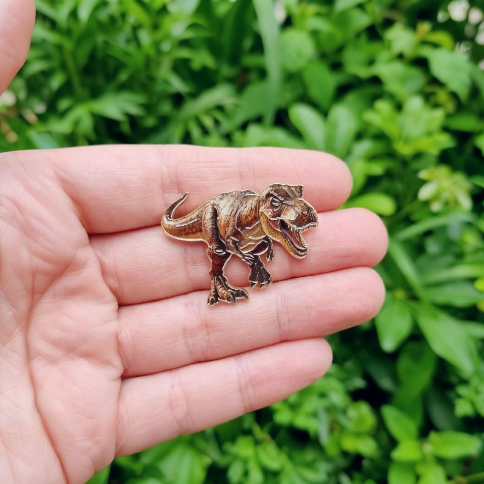 A small T-Rex dinosaur-shaped pin or charm with an antique bronze finish is held on a person's palm, with blurred green foliage in the background.