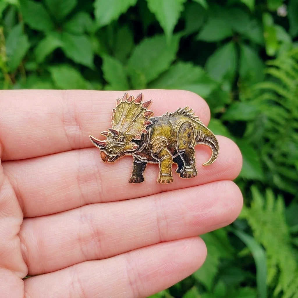 A small triceratops dinosaur pin with an antique gold and bronze finish, held on a person's fingers against a blurred green leaf background.