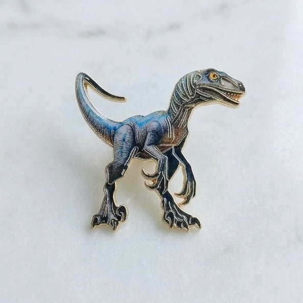 A small dinosaur raptor enamel pin in metallic blue and black with gold-toned details, resting on a white marble surface.
