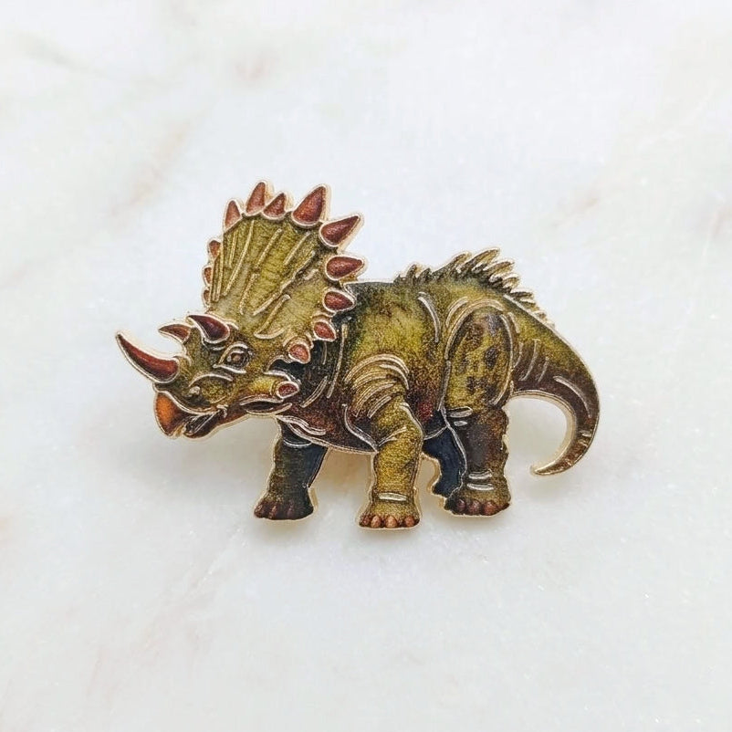 A small dinosaur pin shaped like a Triceratops, with an antique gold finish, resting on a white marble surface.