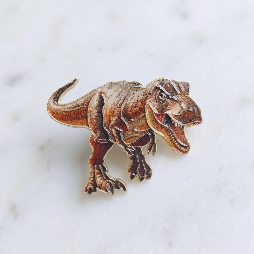 A small dinosaur pin shaped like a brown T-rex, photographed on a light marble surface.