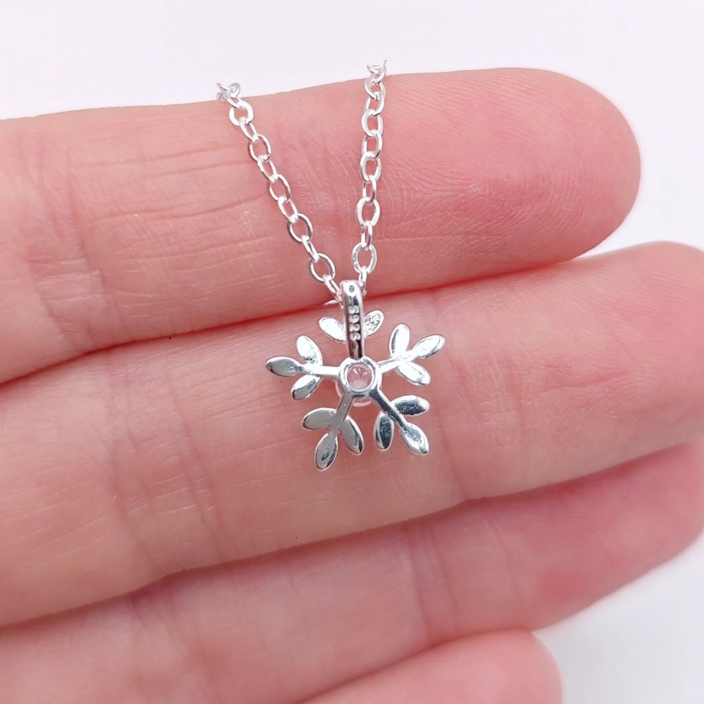 The back of a silver snowflake pendant showing the hallmark S925, held on a person's fingers against a white background.