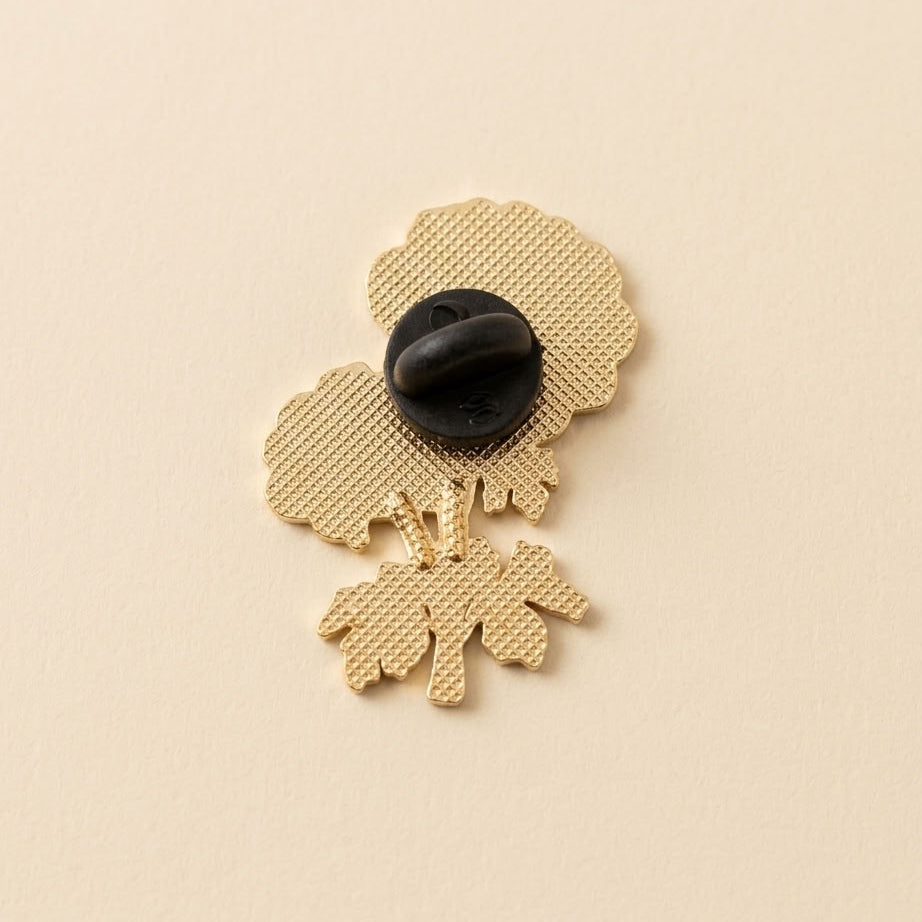 A gold-tone metal flower-shaped pin with a textured pattern, featuring a black plastic clasp on the back, set against a solid light beige background.