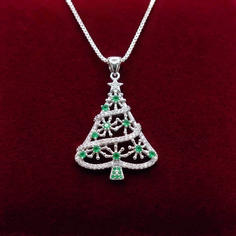 A silver Christmas tree pendant 925 silver necklace with green and clear gemstone accents, resting on a dark red velvet surface with a blurred festive background.