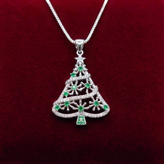 A silver Christmas tree pendant 925 silver necklace with green and clear gemstone accents, resting on a dark red velvet surface with a blurred festive background.