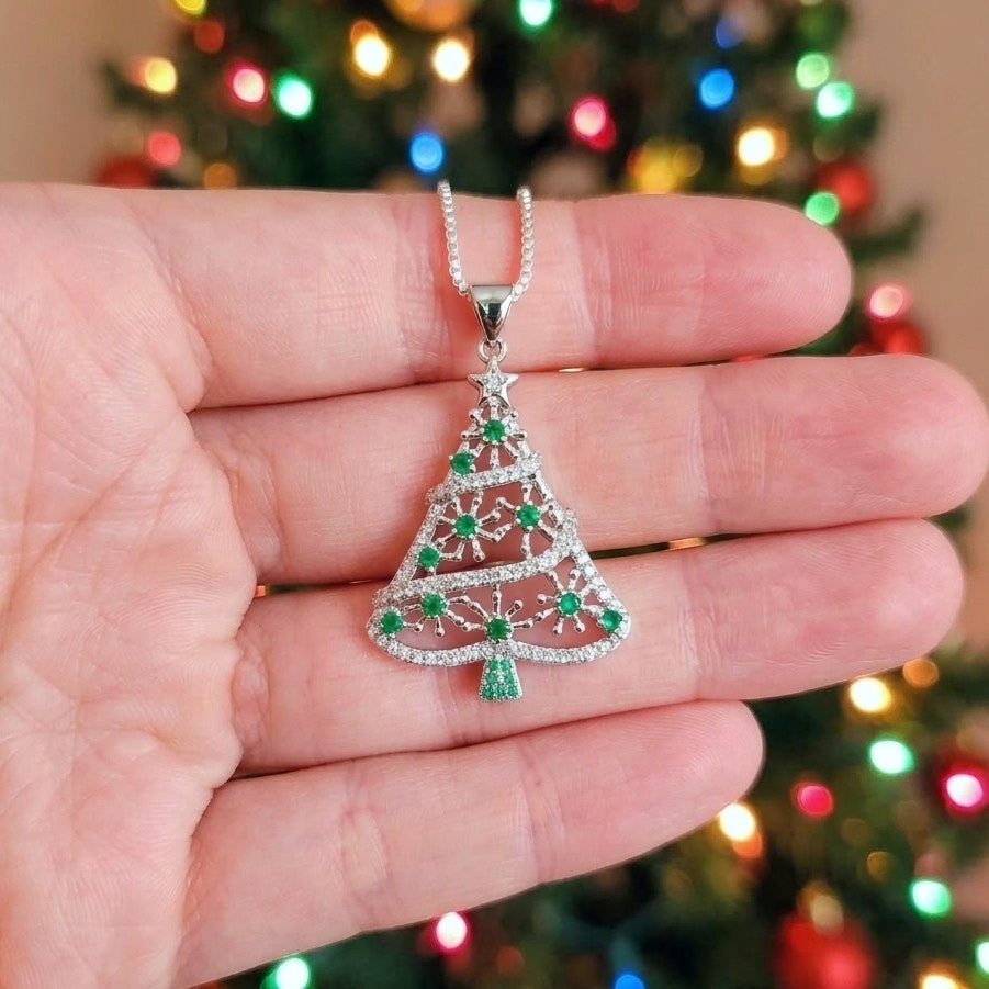 A silver Christmas tree pendant necklace with green cubic zirconia gemstone accents, held in a person's hand against a blurred Christmas tree background with colorful lights.