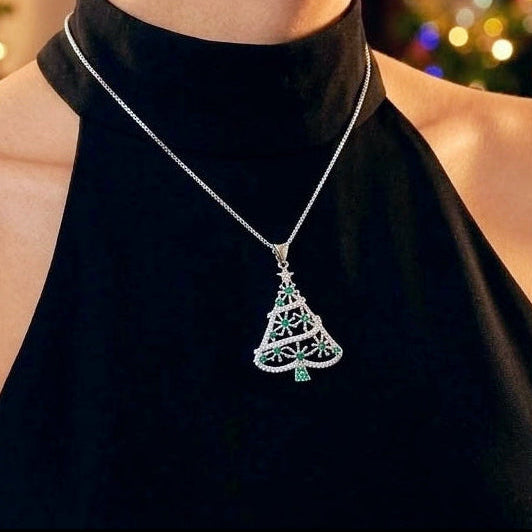 A woman wearing a silver filigree Christmas tree pendant necklace with a small green cubic zirconia accent, paired with a black halter top, set against a festive blurred Christmas tree background.