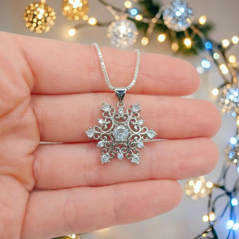 A silver snowflake pendant necklace with clear crystals, held in a person's hand against a festive background of warm and cool holiday string lights.