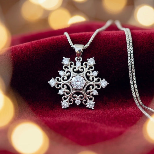 Filigree silver snowflake pendant with bright cubic zirconia crystals on a venetian box chain, resting on dark red velvet material with colourful blurred festive lights in the background.