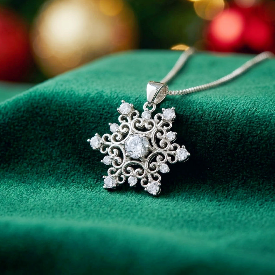 A silver snowflake pendant necklace with clear crystals, resting on a textured dark green velvet fabric with a festive blurred holiday background.