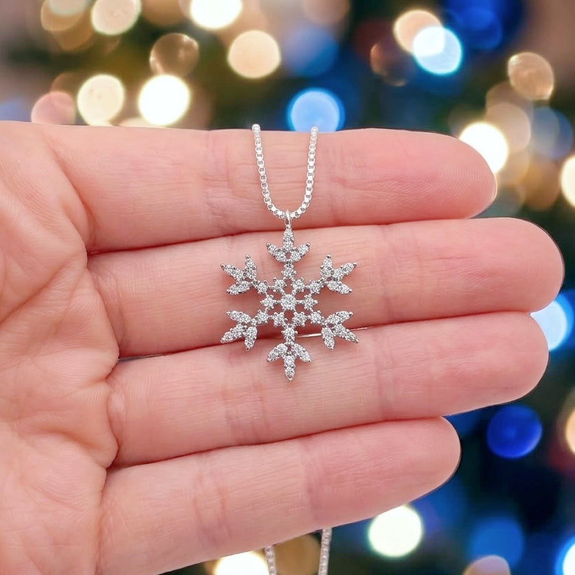 A silver snowflake pendant necklace with clear zirconia stones, held in a person's hand against a blurred festive background with colorful bokeh lights.