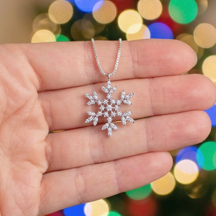 A silver snowflake pendant necklace with clear stones, held in the palm of a hand against a blurred background of colorful Christmas tree lights.