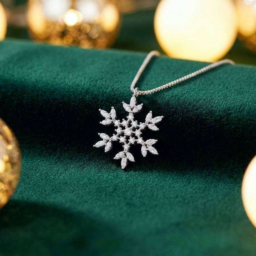 A silver snowflake pendant necklace with sparkling clear stones, resting on a dark green velvet surface with warm golden bokeh lights in the background.