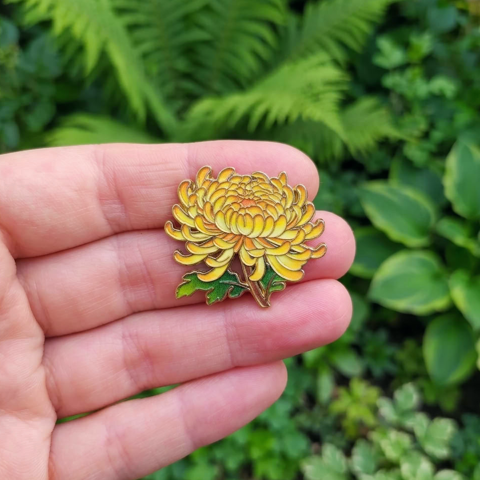 A yellow chrysanthemum flower enamel pin with green leaves, held on a person's fingers against a blurred green garden background.