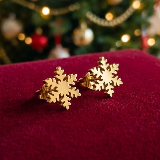 Dainty gold colour snowflake stud earrings made from stainless steel on a red velvet background with a blurred christmas tree in the background.