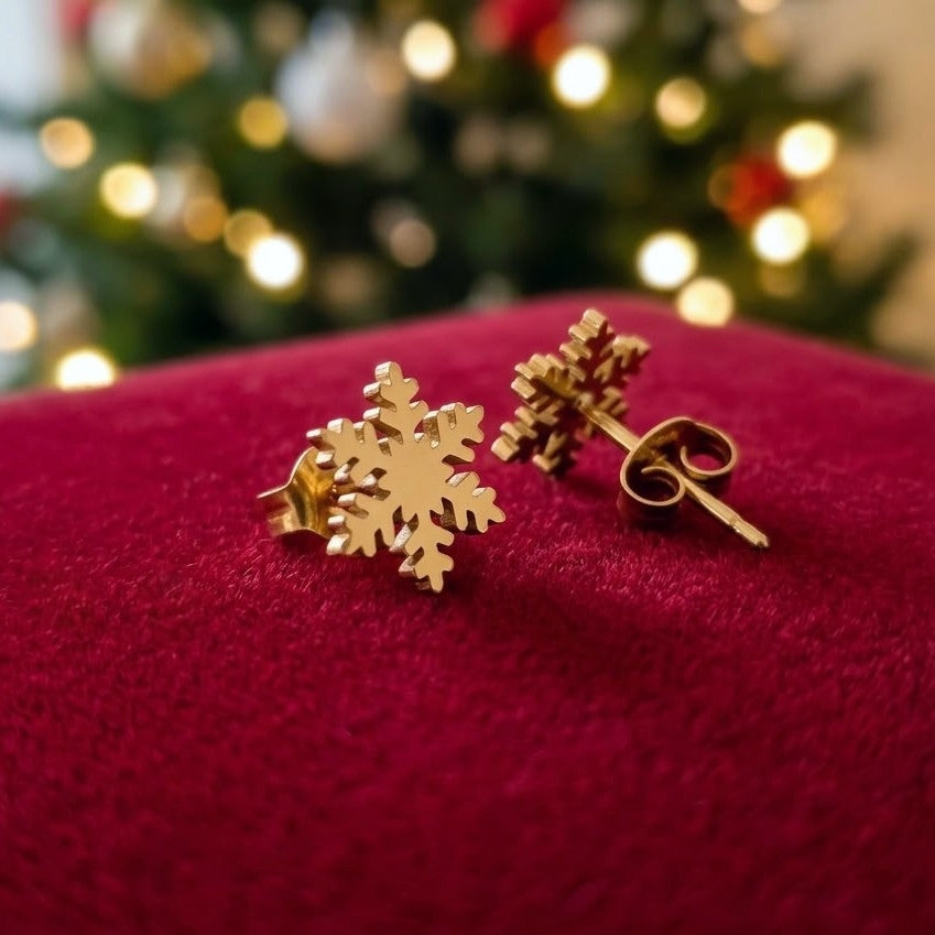 Dainty gold colour snowflake stud earrings made from stainless steel on a red velvet background with a blurred christmas tree in the background.