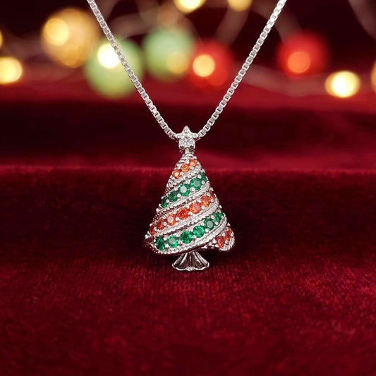 A 925 silver Christmas tree pendant necklace adorned with green and orange cubic zirconia crystals, resting on a deep red velvet surface with a warm bokeh background.