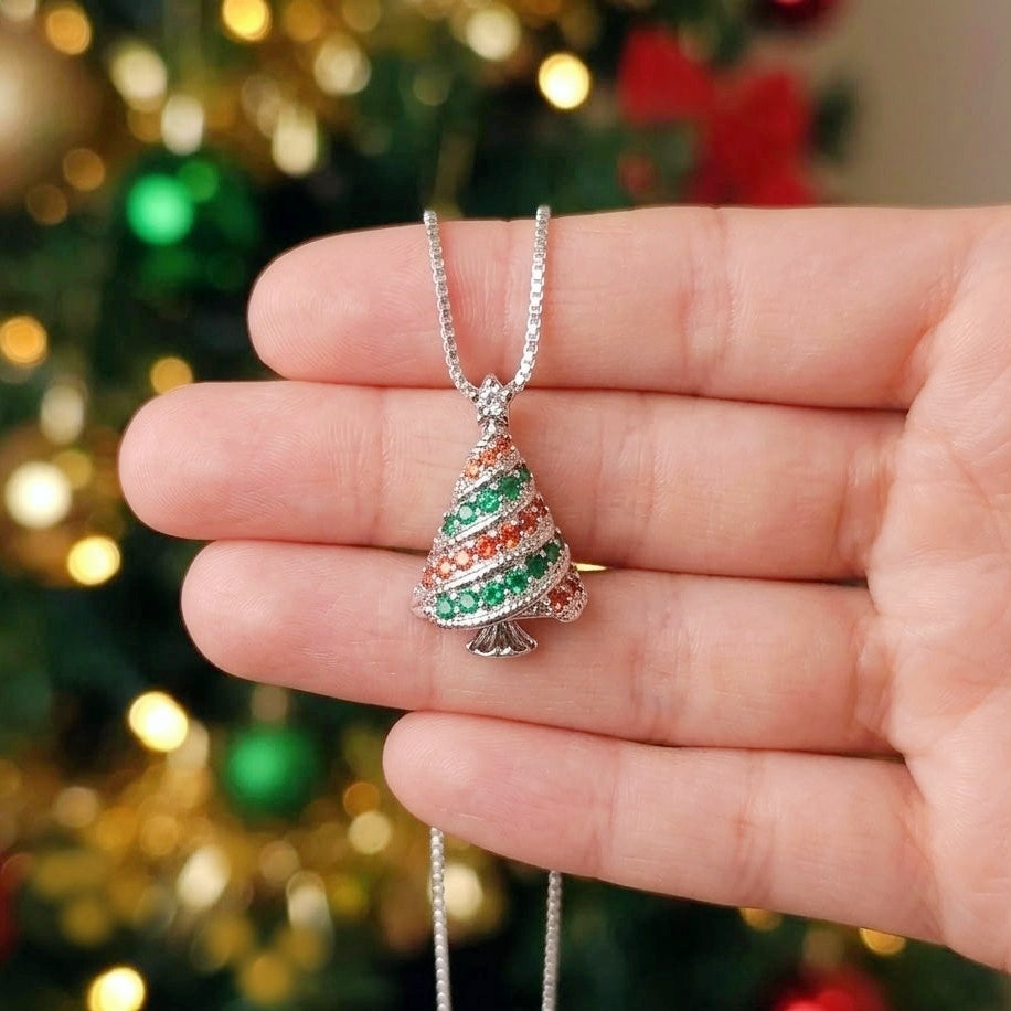 A silver Christmas tree pendant necklace with green and orange striped detailing, held in a person's hand against a blurred Christmas tree background.