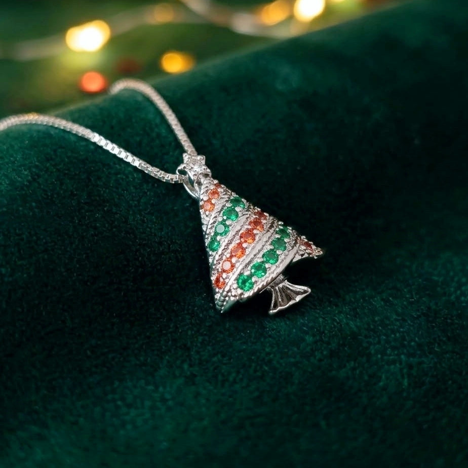 A silver Christmas tree pendant necklace adorned with green and orange rhinestones, resting on a dark green velvet surface with blurred festive lights in the background.