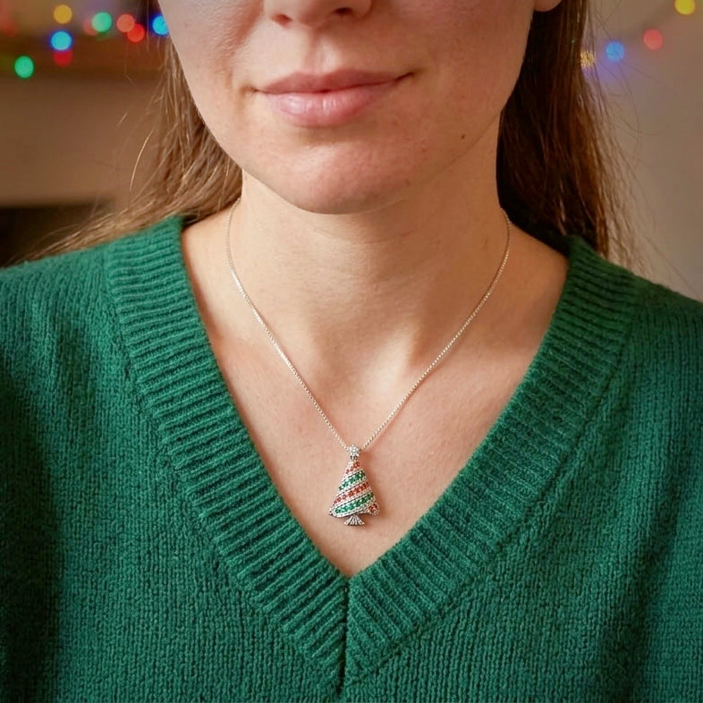 A close-up of a woman wearing a silver necklace with a Christmas tree pendant decorated with otrange, green, and silver stripes, paired with a green V-neck sweater.