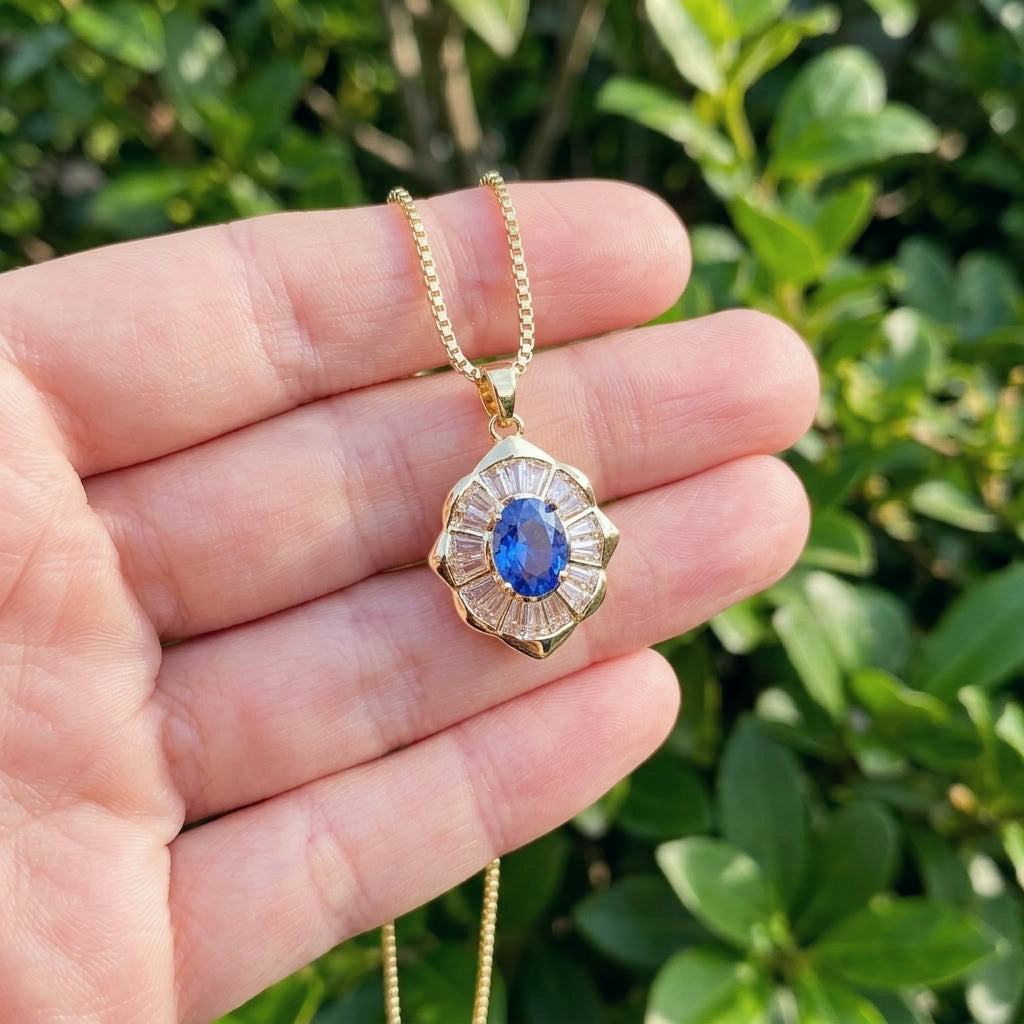 A gold pendant necklace with a blue oval center stone and surrounding clear stones, held in a person's hand against a blurred green outdoor background.