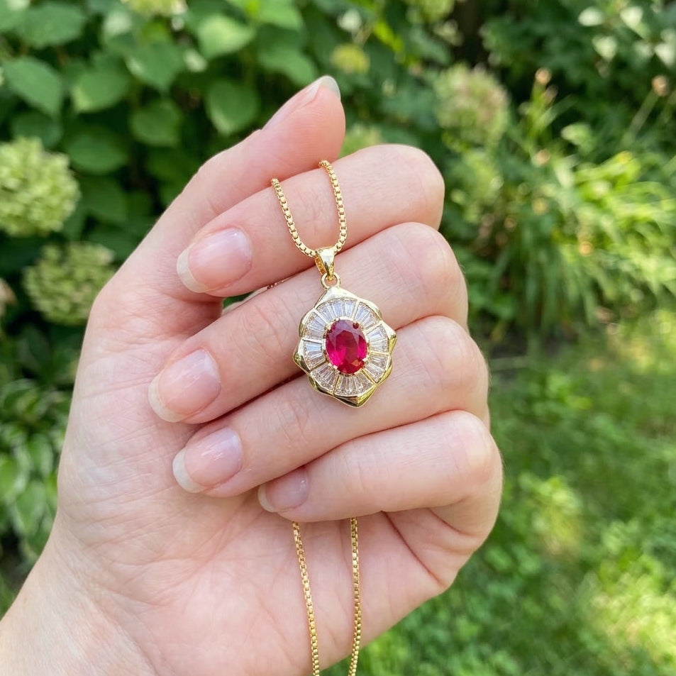 A hand holding a gold pendant necklace with a central round red gemstone and surrounding clear stones, set against a blurred green garden background.