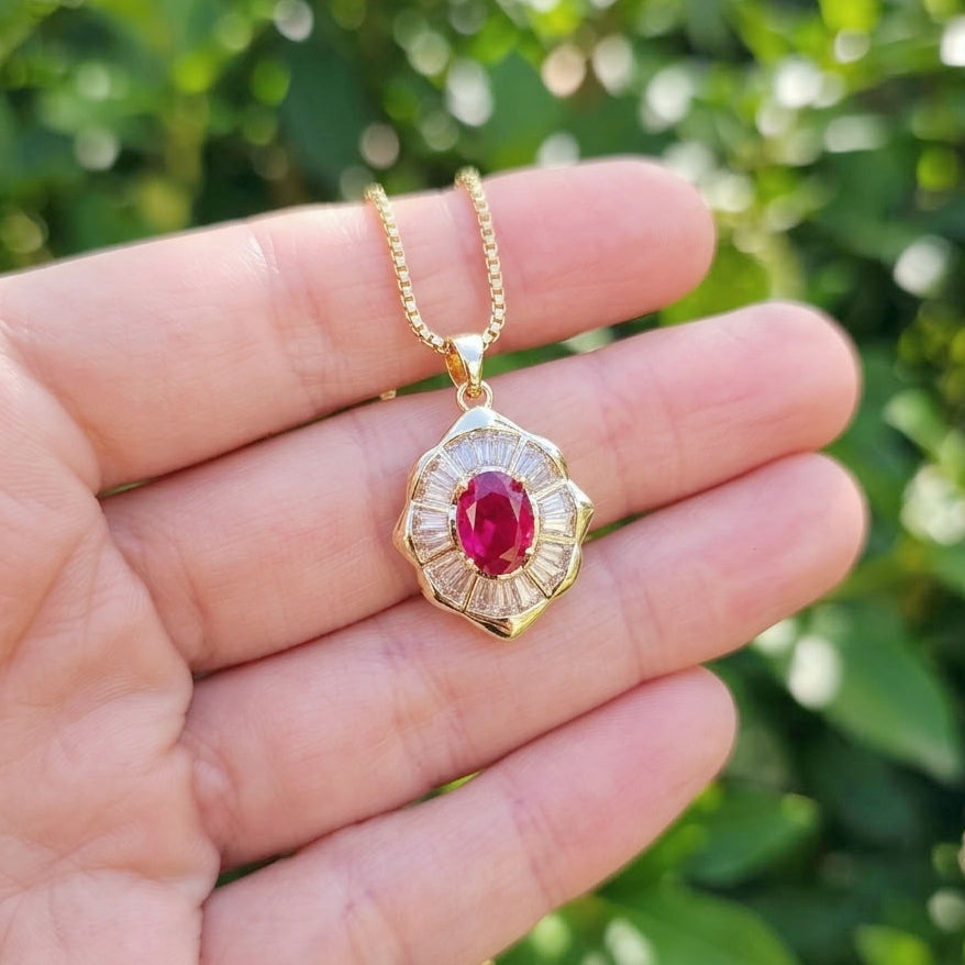 A gold pendant necklace with a central red gemstone and clear accent stones, held in a person's hand against a blurred green outdoor background.