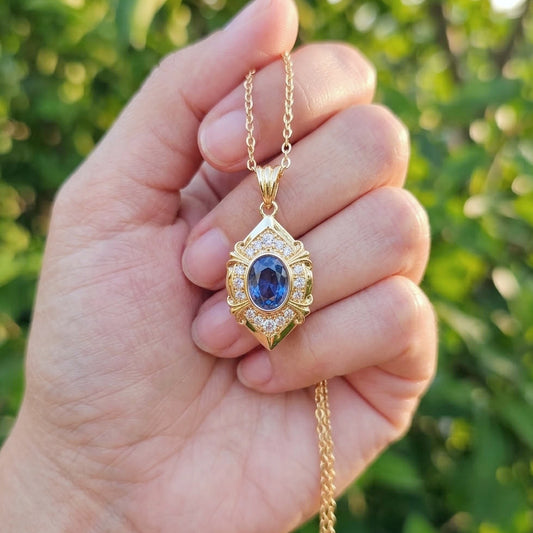 A gold pendant necklace with a blue oval gemstone and small clear stones, held in a person's hand against a blurred green outdoor background.