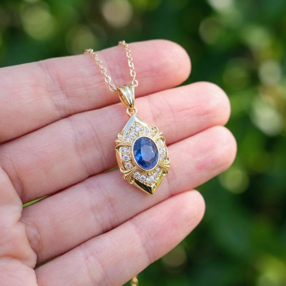 A gold pendant necklace with a central oval blue sapphire and small clear accent stones, held on a person's fingers against a blurred green outdoor background.