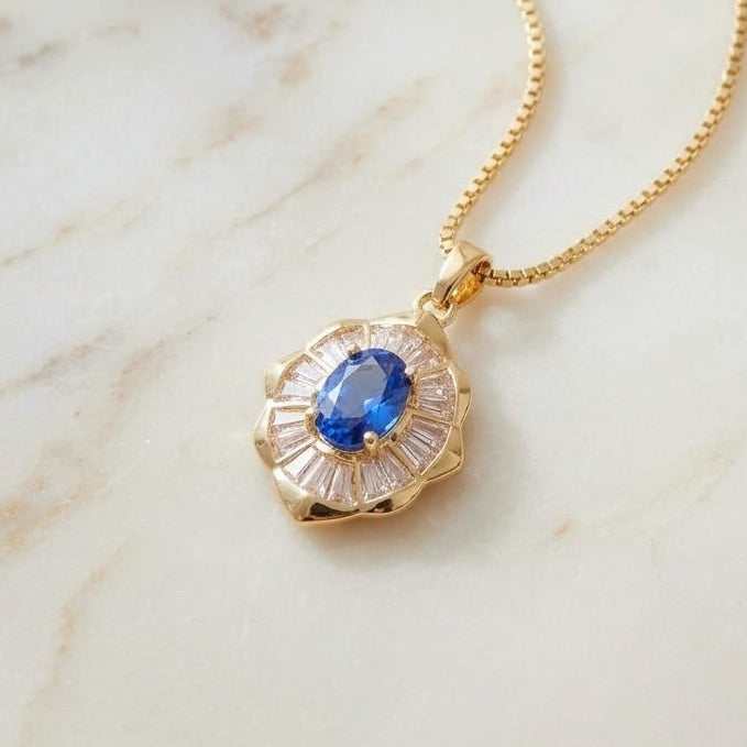 A gold pendant necklace featuring a central oval blue gemstone surrounded by a halo of small clear stones, resting on a light marble surface.